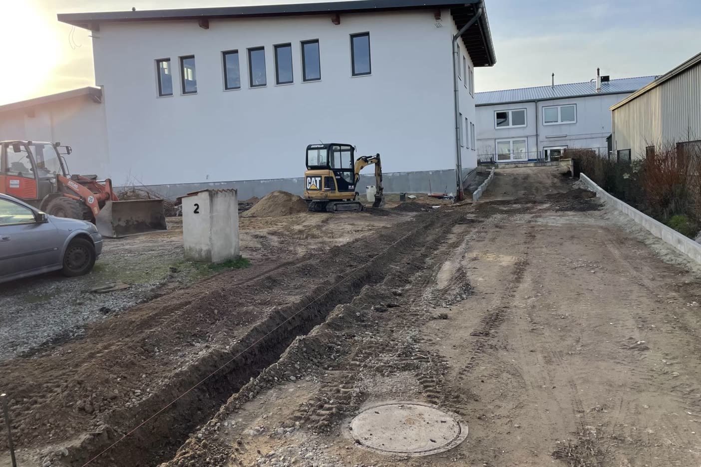 Zufahrt am Gewerbegebäude vor den Arbeiten: Offener Leitungsgraben im Erdplanum, Minibagger, gesetzter Kontrollschacht
